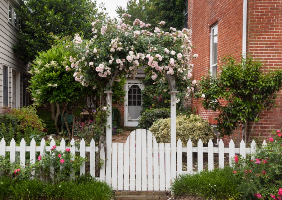 Front entry arbor with gate topped with colorful flowers, and a white picket fence